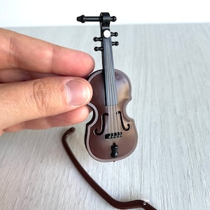 May include: A miniature brown violin with black strings and a black stand. The violin has a small black knob at the bottom.
