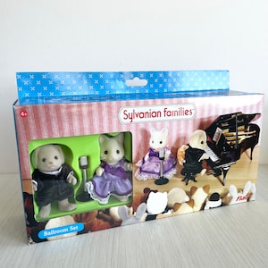 Puede incluir: Juego de Sylvanian Families Ballroom Set con tres figuras, un perro blanco, un gato blanco y un gato blanco con un vestido morado. El juego incluye un micrófono y un piano de cola negro. La caja tiene un fondo azul y blanco con el texto "Sylvanian Families" y "Ballroom Set".
