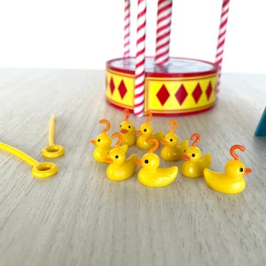Op de afbeelding: Een set gele rubberen eendjes met oranje snavels en haken op hun kop. De eendjes zijn in een rij op een licht houten oppervlak gerangschikt. Er is een rood-wit gestreepte draaimolen op de achtergrond.