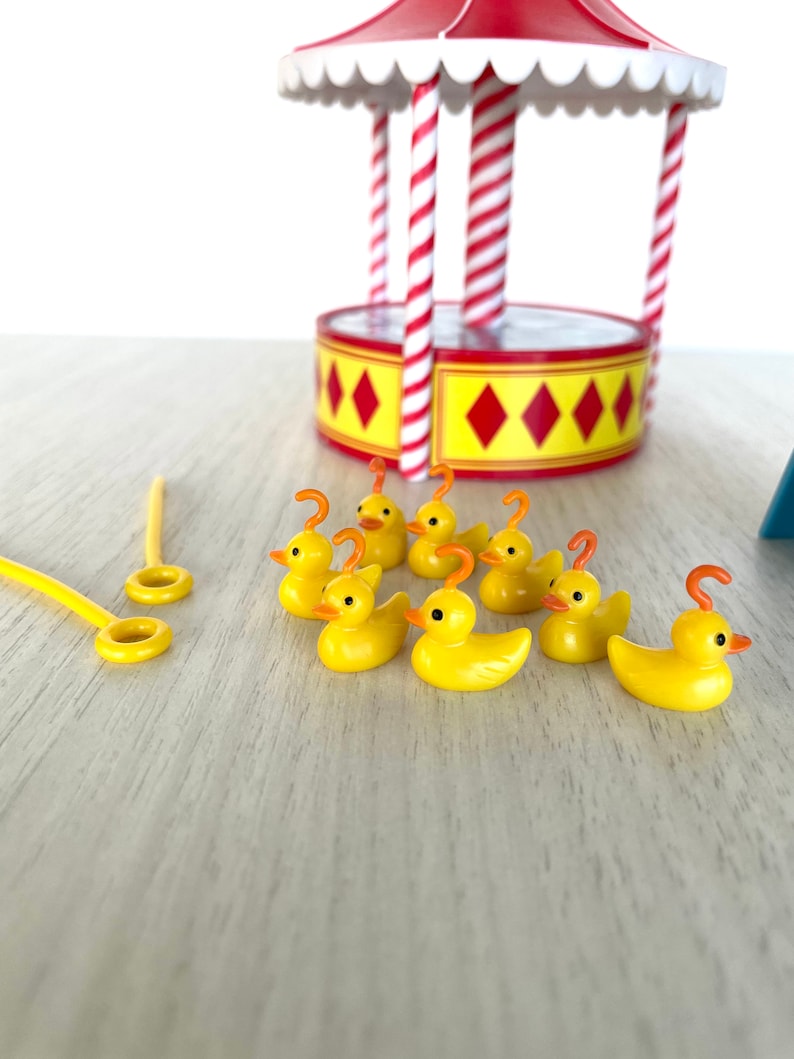 Op de afbeelding: Een set gele rubberen eendjes met oranje snavels en haken op hun kop. De eendjes zijn in een rij op een licht houten oppervlak gerangschikt. Er is een rood-wit gestreepte draaimolen op de achtergrond.