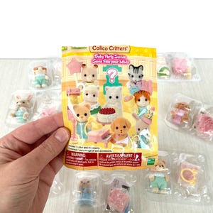 Peut inclure: Un sac en plastique contenant un ensemble Calico Critters Baby Party Series. L'ensemble comprend une variété de petites figurines, dont un chat, un chien, un lapin et un ours. Le sac comprend également un piano rose et un gâteau avec des bougies. Le sac est étiqueté "Calico Critters Baby Party Series" et "Série Fête pour bébés".