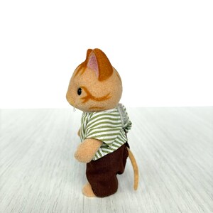 May include: A small, orange and tan cat figurine wearing a green and white striped shirt and brown pants. The cat has a long, tan tail and is standing on a light-colored surface. The figurine is a toy.