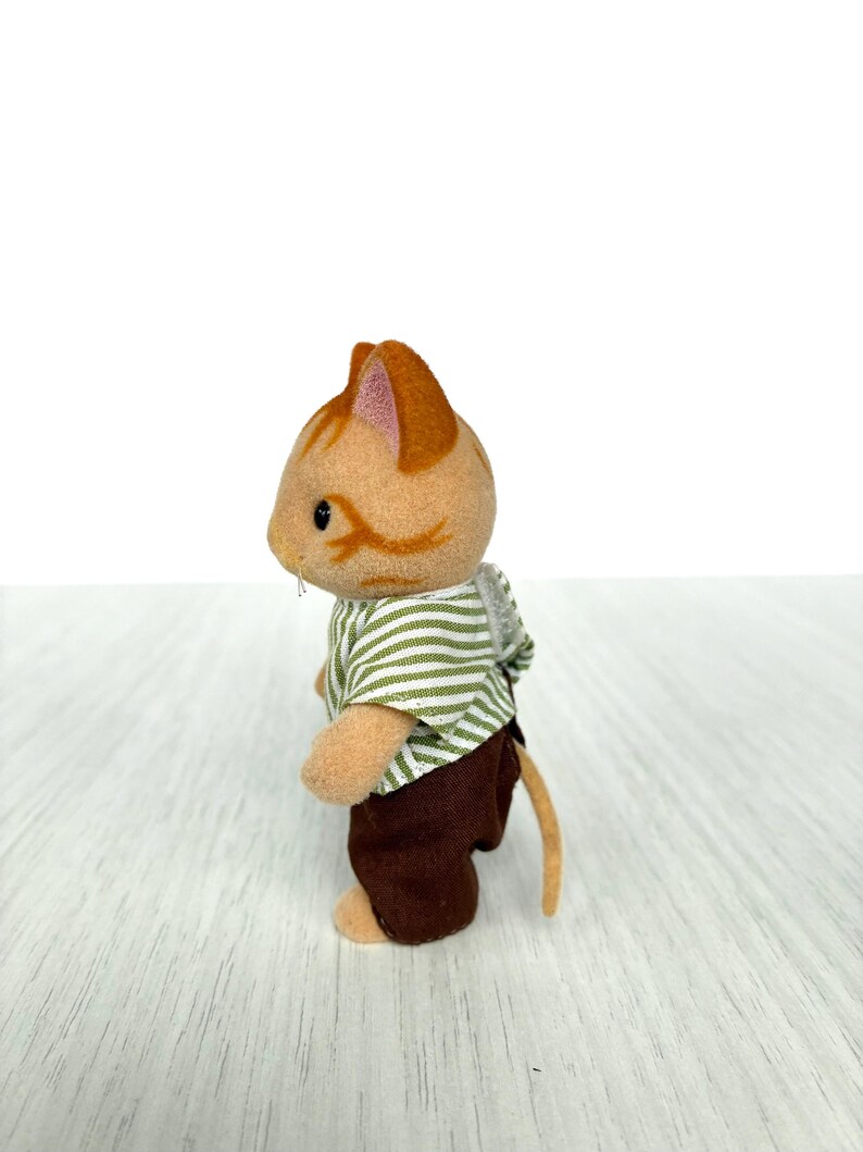 May include: A small, orange and tan cat figurine wearing a green and white striped shirt and brown pants. The cat has a long, tan tail and is standing on a light-colored surface. The figurine is a toy.