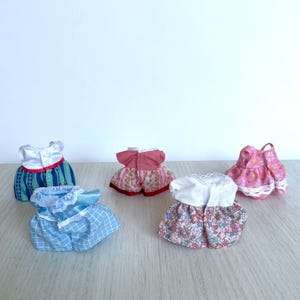 May include: Six miniature doll dresses in various colors and patterns, including pink, blue, and white. The dresses are made of fabric and have lace trim.
