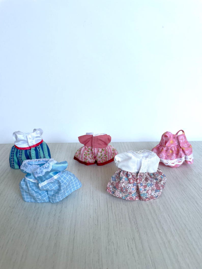 May include: Six miniature doll dresses in various colors and patterns, including pink, blue, and white. The dresses are made of fabric and have lace trim.