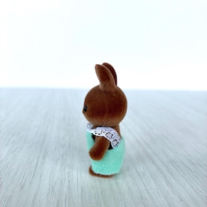 May include: A brown rabbit figurine wearing a light green outfit with a white lace collar. The toy rabbit has small black eyes and is standing on a light-colored surface.