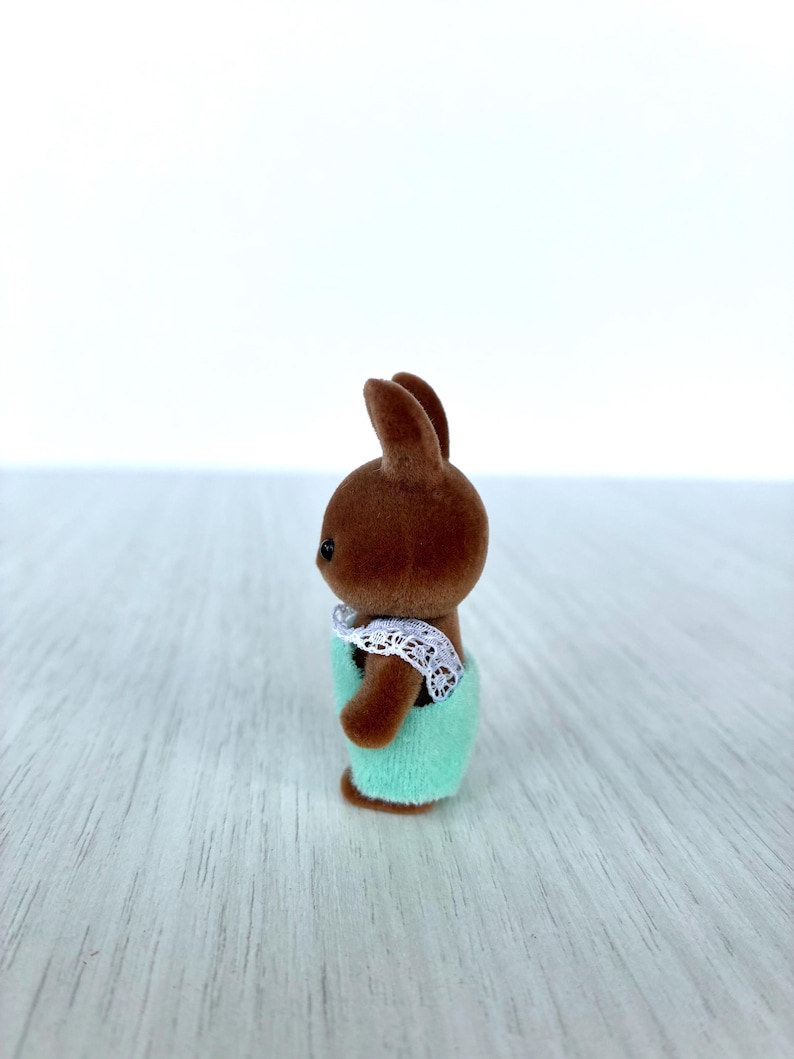 May include: A brown rabbit figurine wearing a light green outfit with a white lace collar. The toy rabbit has small black eyes and is standing on a light-colored surface.