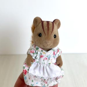 May include: A small brown squirrel figurine wearing a white and floral dress with a white apron. The dress has a lace trim.