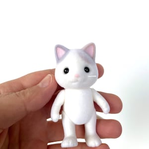 May include: A white plush cat toy with gray ears and black eyes. The cat is standing on its four paws and has a pink nose.