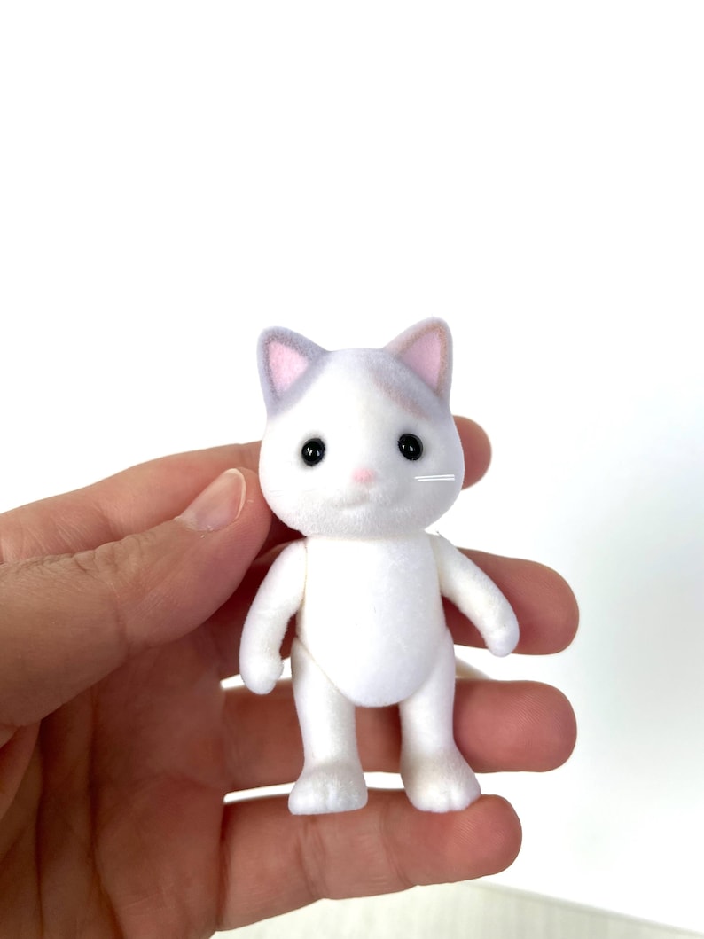 May include: A white plush cat toy with gray ears and black eyes. The cat is standing on its four paws and has a pink nose.