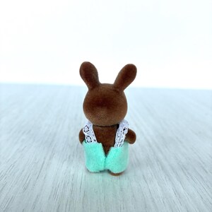 May include: A small brown rabbit figurine wearing a light green jumpsuit with white lace trim. The toy rabbit has long ears and is standing on a light-colored surface. The figurine is a miniature toy.