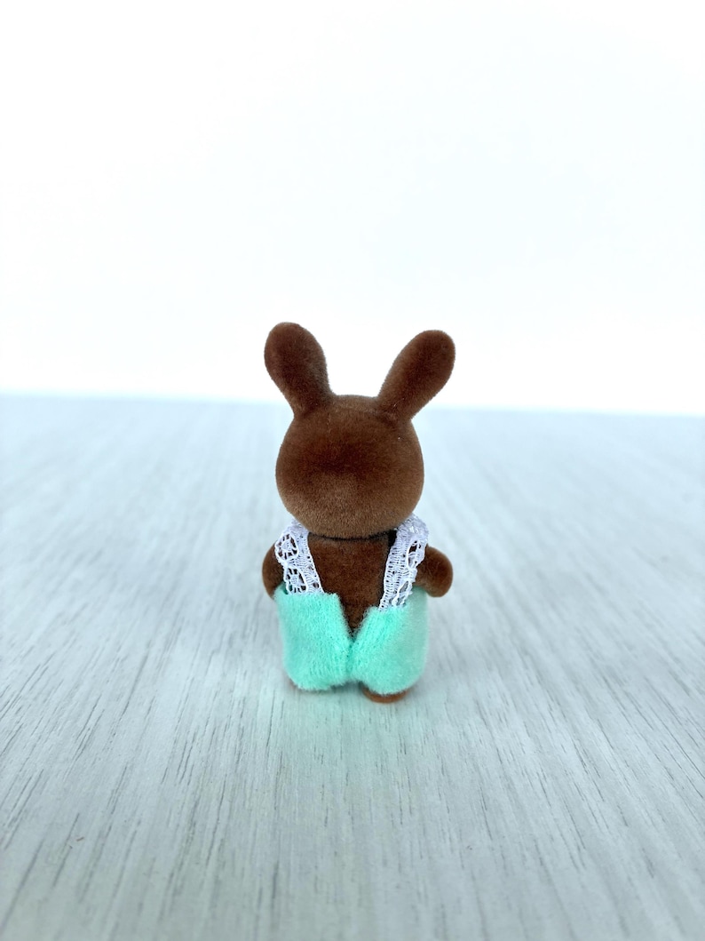 May include: A small brown rabbit figurine wearing a light green jumpsuit with white lace trim. The toy rabbit has long ears and is standing on a light-colored surface. The figurine is a miniature toy.