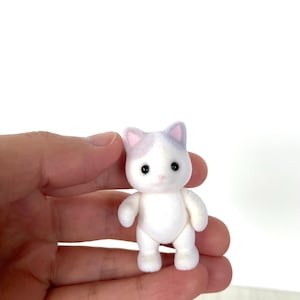 May include: A small white cat figurine with purple ears and black eyes. The figurine is standing on its four paws and has a fluffy texture.
