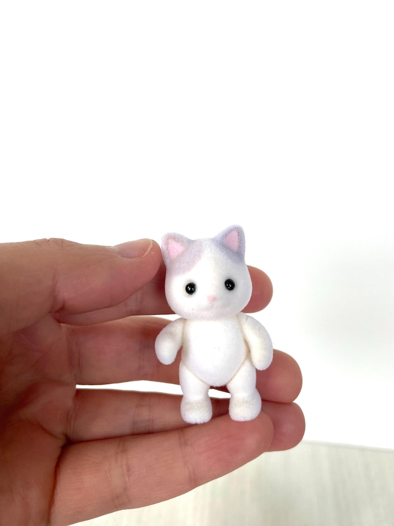 May include: A small white cat figurine with purple ears and black eyes. The figurine is standing on its four paws and has a fluffy texture.