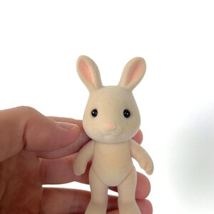 May include: A small, cream-colored rabbit figurine with pink ears and black eyes. The figurine is standing upright with its feet together.