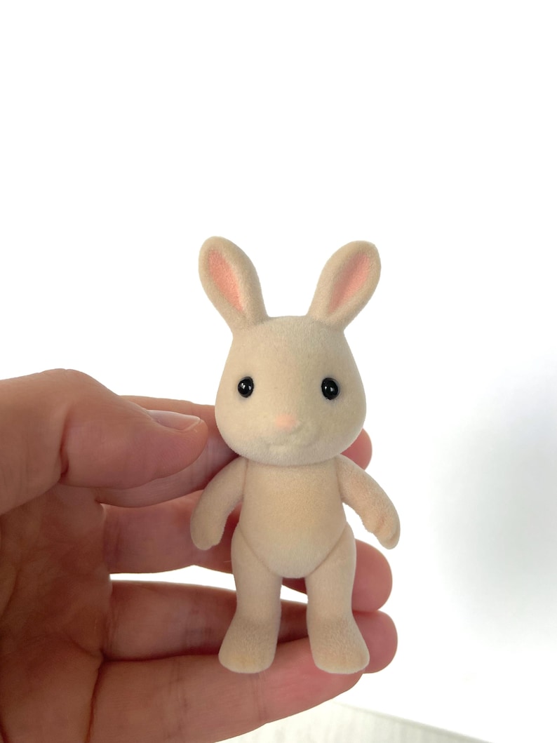 May include: A small, cream-colored rabbit figurine with pink ears and black eyes. The figurine is standing upright with its feet together.