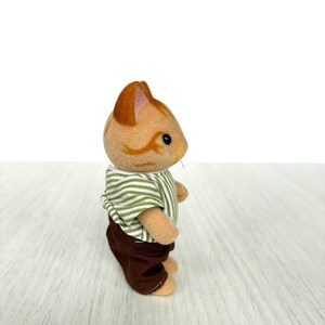 May include: A small, tan cat figurine with brown stripes and a striped green and white shirt. The cat is wearing brown pants. The figurine is standing on a light-colored wooden surface.