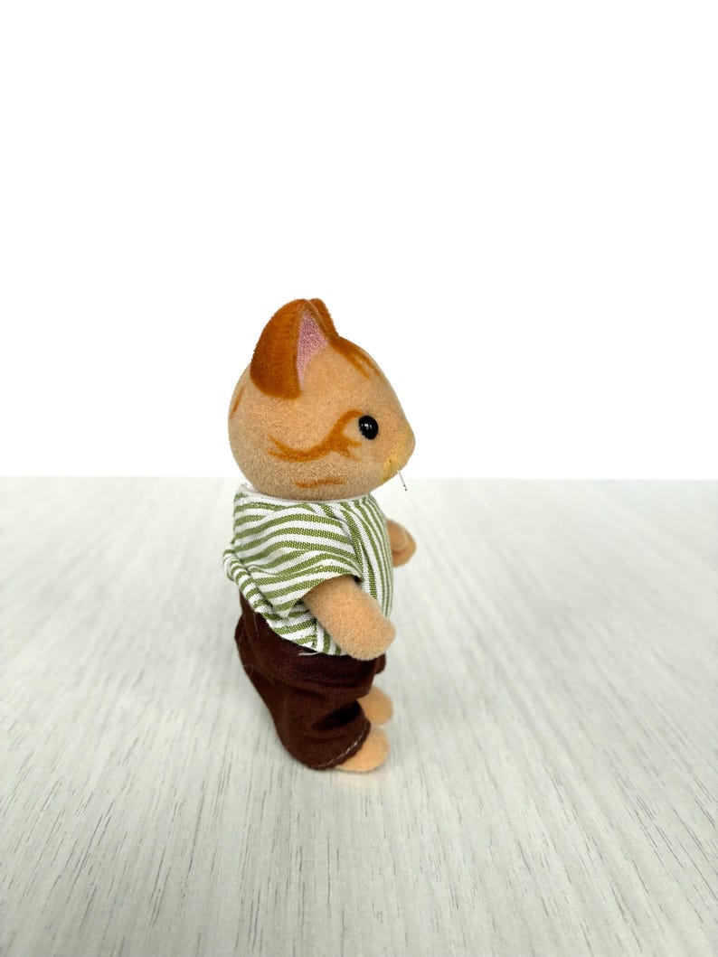 May include: A small, tan cat figurine with brown stripes and a striped green and white shirt. The cat is wearing brown pants. The figurine is standing on a light-colored wooden surface.