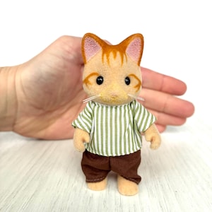 May include: A miniature orange tabby cat figurine wearing a green and white striped shirt and brown pants. The cat has black eyes and white whiskers. The figurine is standing on a white wooden surface.