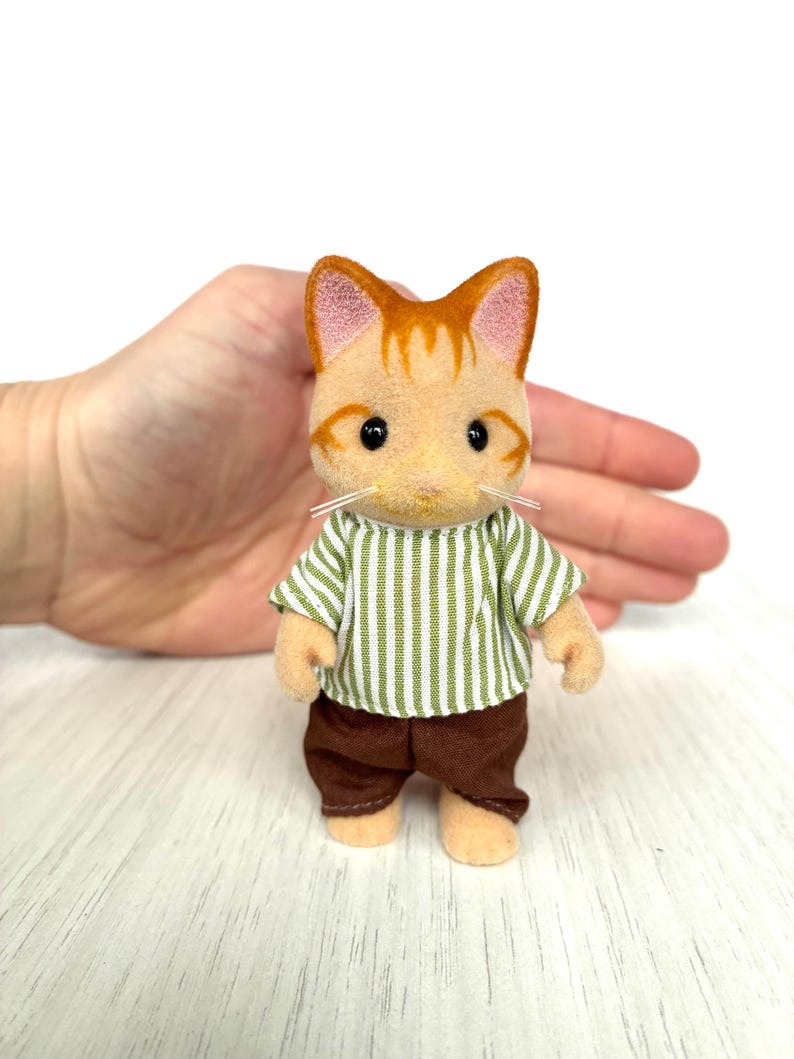 May include: A miniature orange tabby cat figurine wearing a green and white striped shirt and brown pants. The cat has black eyes and white whiskers. The figurine is standing on a white wooden surface.