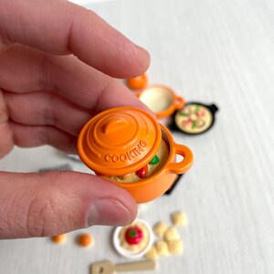 May include: Orange miniature cooking pot with a lid and a handle. The pot is filled with a simulated food dish. The pot has the word "COOKING" printed on the side.