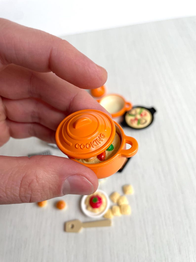 May include: Orange miniature cooking pot with a lid and a handle. The pot is filled with a simulated food dish. The pot has the word "COOKING" printed on the side.