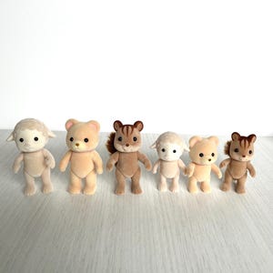 Peut inclure: Six petites figurines d'animaux en peluche, dont un agneau, un ours, deux écureuils et deux autres agneaux. Les figurines sont toutes debout sur une surface en bois clair.