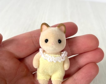 May include: A small, tan-colored cat figurine wearing a yellow jumpsuit with a white lace collar. The cat has brown-tipped ears and black eyes. The figurine is a miniature toy, likely for a dollhouse or a collector's item.