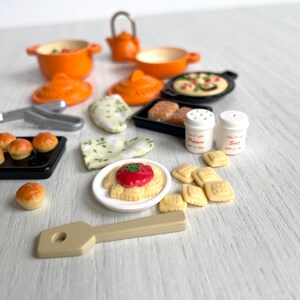 May include: A miniature kitchen playset with orange pots, pans, and a teapot. There are also miniature food items, including bread rolls, pizza, and pasta. The set also includes a wooden spatula and two salt and pepper shakers.