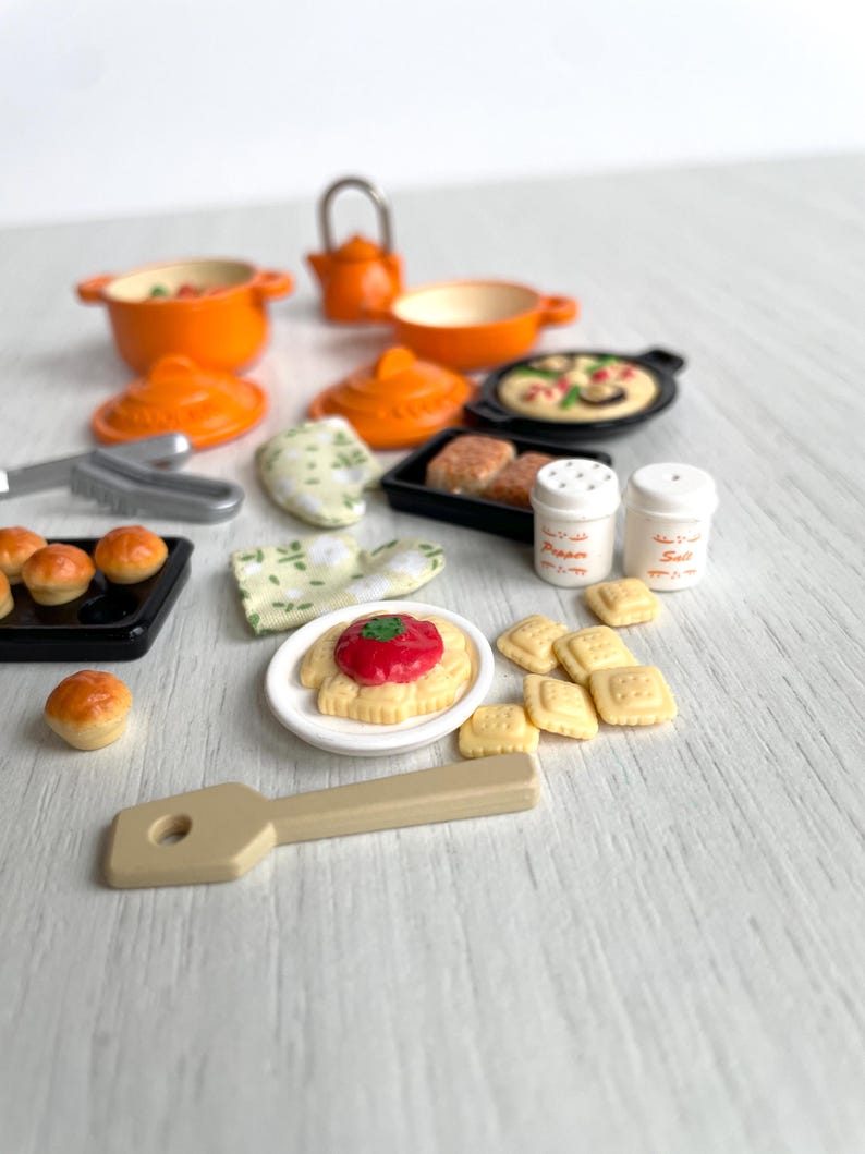 May include: A miniature kitchen playset with orange pots, pans, and a teapot. There are also miniature food items, including bread rolls, pizza, and pasta. The set also includes a wooden spatula and two salt and pepper shakers.