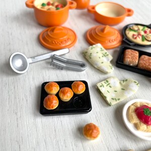 May include: A miniature kitchen playset with orange pots, pans, and lids, a silver ladle and tongs, a baking sheet with four small bread rolls, a white plate with a red sauce and pasta, a wooden spatula, and two green and white dish towels.