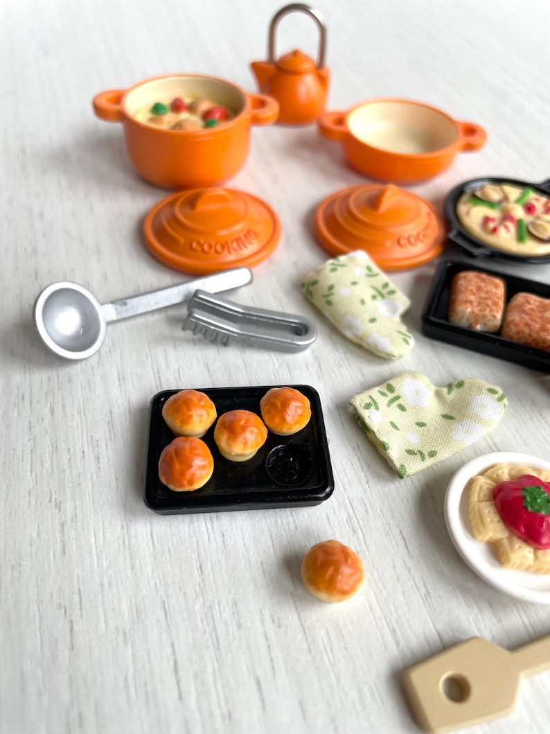 May include: A miniature kitchen playset with orange pots, pans, and lids, a silver ladle and tongs, a baking sheet with four small bread rolls, a white plate with a red sauce and pasta, a wooden spatula, and two green and white dish towels.