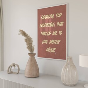 May include: A framed print with a red background and gold lettering that reads "Grateful for everything that forced me to love myself more". The print is hanging on a white wall above a wooden dresser with a vase of dried pampas grass and a decorative object.