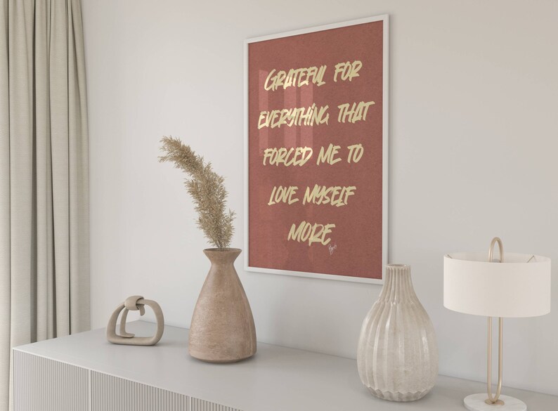 May include: A framed print with a red background and gold lettering that reads "Grateful for everything that forced me to love myself more". The print is hanging on a white wall above a wooden dresser with a vase of dried pampas grass and a decorative object.