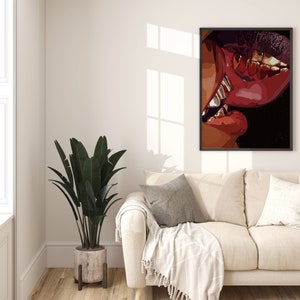 May include: A framed art print featuring a close-up of two sets of lips in shades of red and brown, with gold teeth visible. The artwork is displayed on a white wall above a beige sofa, with a large potted plant in the corner.