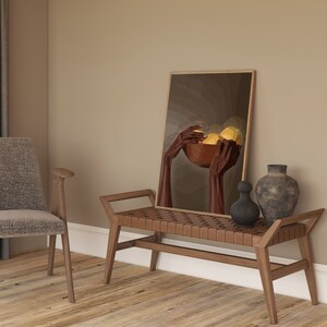May include: A wooden bench with a woven leather seat sits in a room with a brown wall. A framed painting of hands holding a bowl of fruit hangs on the wall above the bench. A vase and a decorative object sit on the bench.