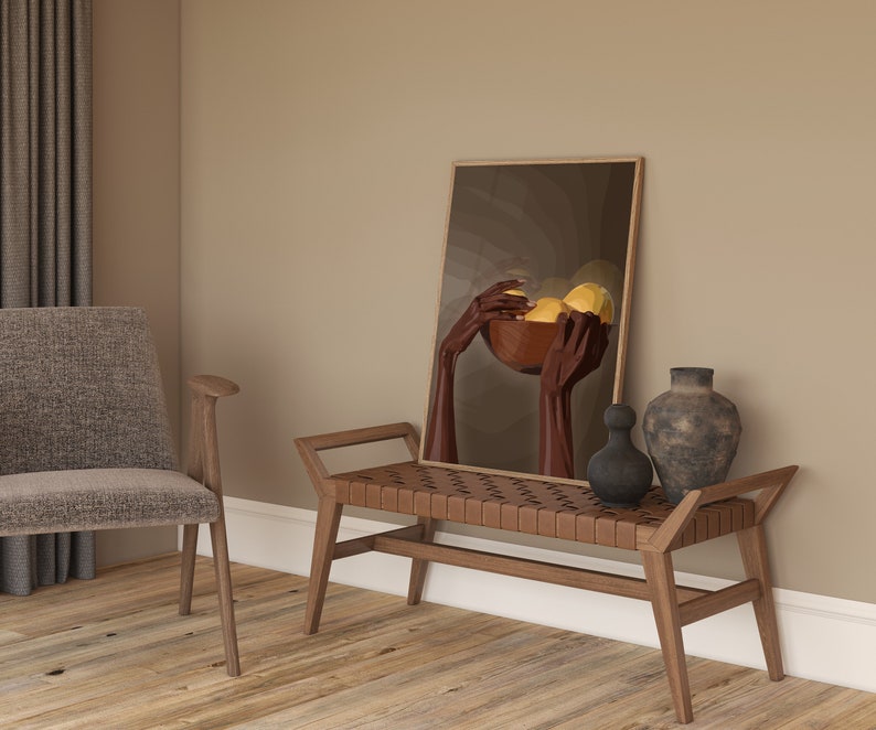 May include: A wooden bench with a woven leather seat sits in a room with a brown wall. A framed painting of hands holding a bowl of fruit hangs on the wall above the bench. A vase and a decorative object sit on the bench.