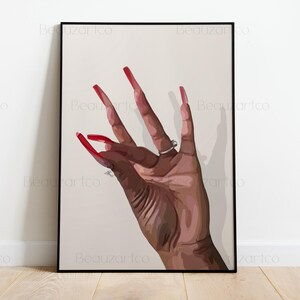 Peut inclure: Impression artistique encadrée représentant une main avec de longs ongles rouges. La main porte deux bagues argentées. L'œuvre est posée sur un fond neutre, créant un contraste saisissant avec la peau foncée de la main.