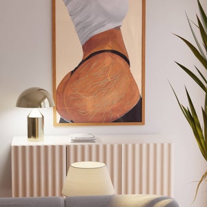 May include: A framed painting of a person's back with a white shirt and black underwear. The painting is in a wooden frame and the background is a light beige color.