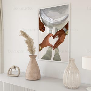 May include: A framed art print depicts two hands forming a heart shape. The hands are dark-skinned, with one wearing a gold bracelet and the other a watch. The artwork is displayed on a white wall, with decorative vases and a lamp in the foreground.