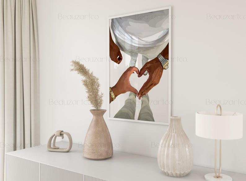 May include: A framed art print depicts two hands forming a heart shape. The hands are dark-skinned, with one wearing a gold bracelet and the other a watch. The artwork is displayed on a white wall, with decorative vases and a lamp in the foreground.
