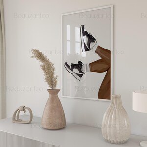 May include: A framed art print featuring an illustration of legs wearing black and white sneakers. The artwork is displayed on a white wall above a white cabinet. Decorative vases and a lamp are on the cabinet. The artwork is a modern, minimalist style.