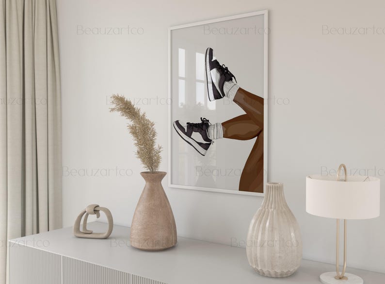 May include: A framed art print featuring an illustration of legs wearing black and white sneakers. The artwork is displayed on a white wall above a white cabinet. Decorative vases and a lamp are on the cabinet. The artwork is a modern, minimalist style.
