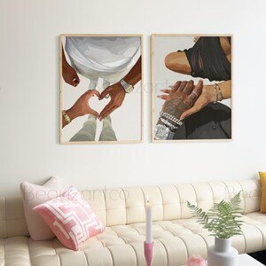 May include: Two framed art prints depicting hands forming a heart shape and holding each other. The artwork features neutral tones, with gold bracelets and a watch visible. The prints are displayed above a cream-colored sofa.
