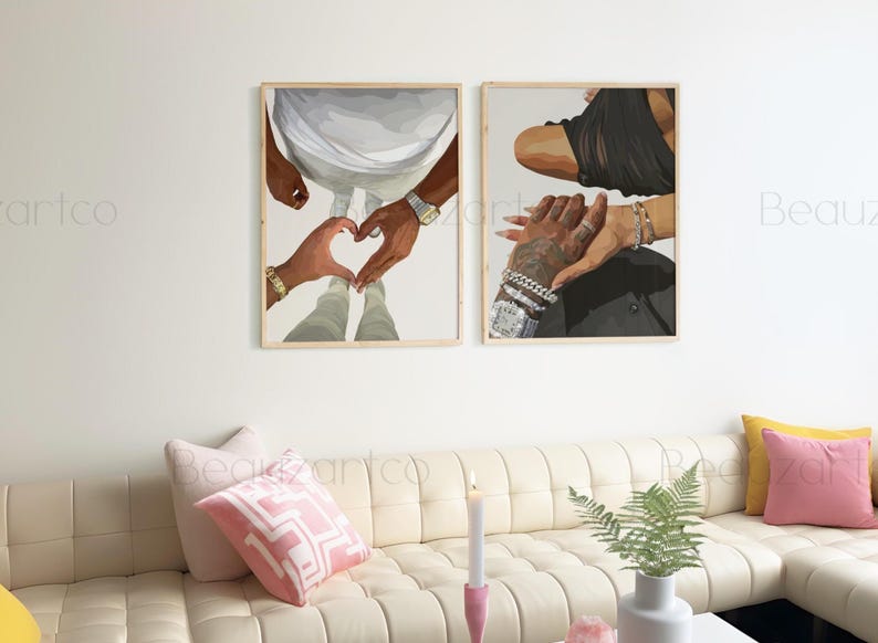 May include: Two framed art prints depicting hands forming a heart shape and holding each other. The artwork features neutral tones, with gold bracelets and a watch visible. The prints are displayed above a cream-colored sofa.