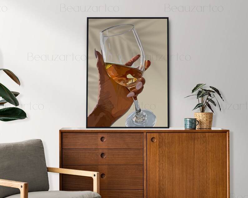 May include: A hand holding a glass of amber-colored liquid. The glass is clear and has a long stem. The hand is brown and has long, manicured nails. The background is a light beige color.