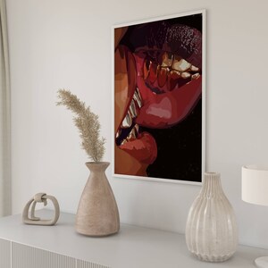 May include: A framed art print featuring a close-up of red lips with gold teeth. The artwork is displayed on a white wall above a white cabinet, next to a vase with dried plants and a decorative lamp.