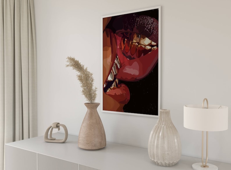 May include: A framed art print featuring a close-up of red lips with gold teeth. The artwork is displayed on a white wall above a white cabinet, next to a vase with dried plants and a decorative lamp.