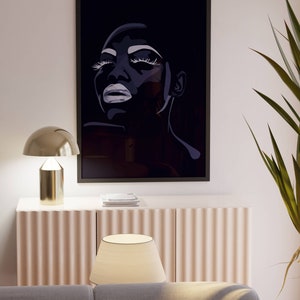May include: A black and white framed print of a woman's face with dramatic makeup. The print is hanging on a white wall above a light-colored cabinet with a lamp on top.