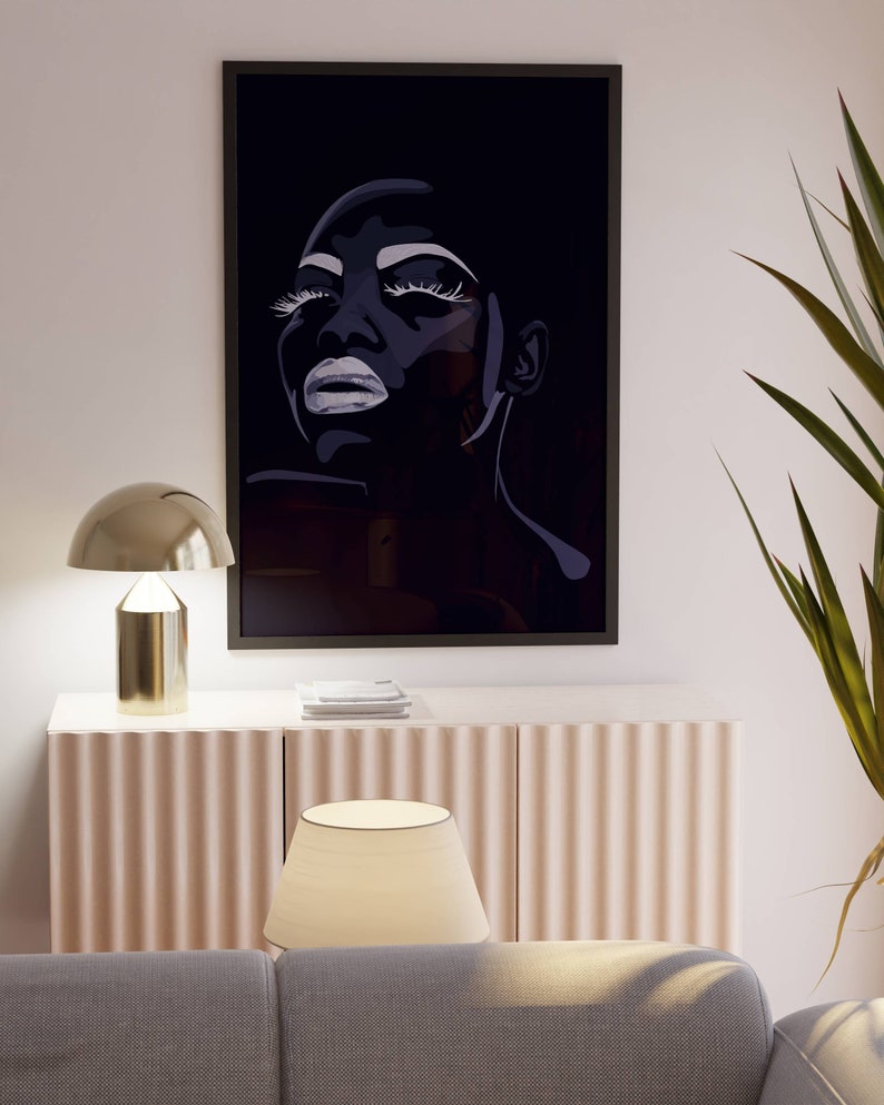 May include: A black and white framed print of a woman's face with dramatic makeup. The print is hanging on a white wall above a light-colored cabinet with a lamp on top.